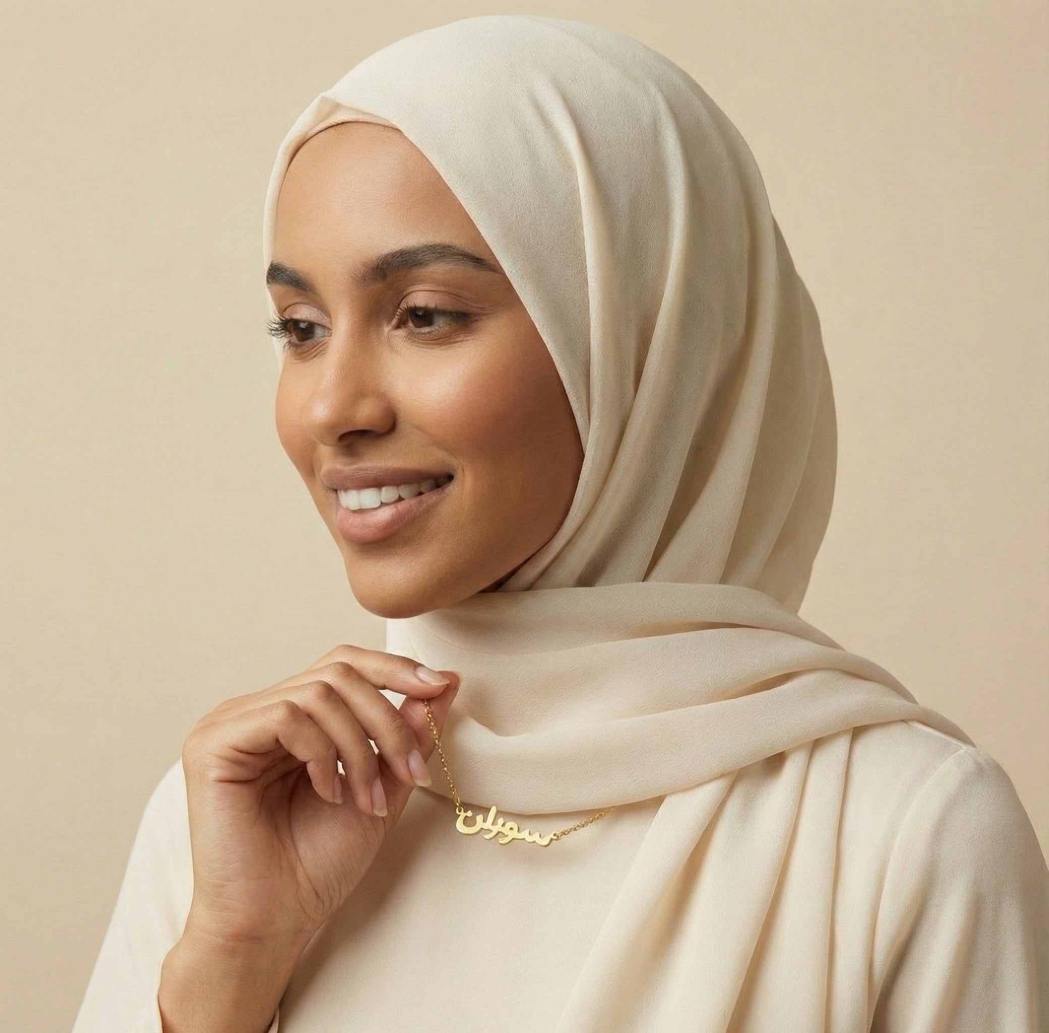 Woman wearing a beige hijab and gold necklace against a beige background