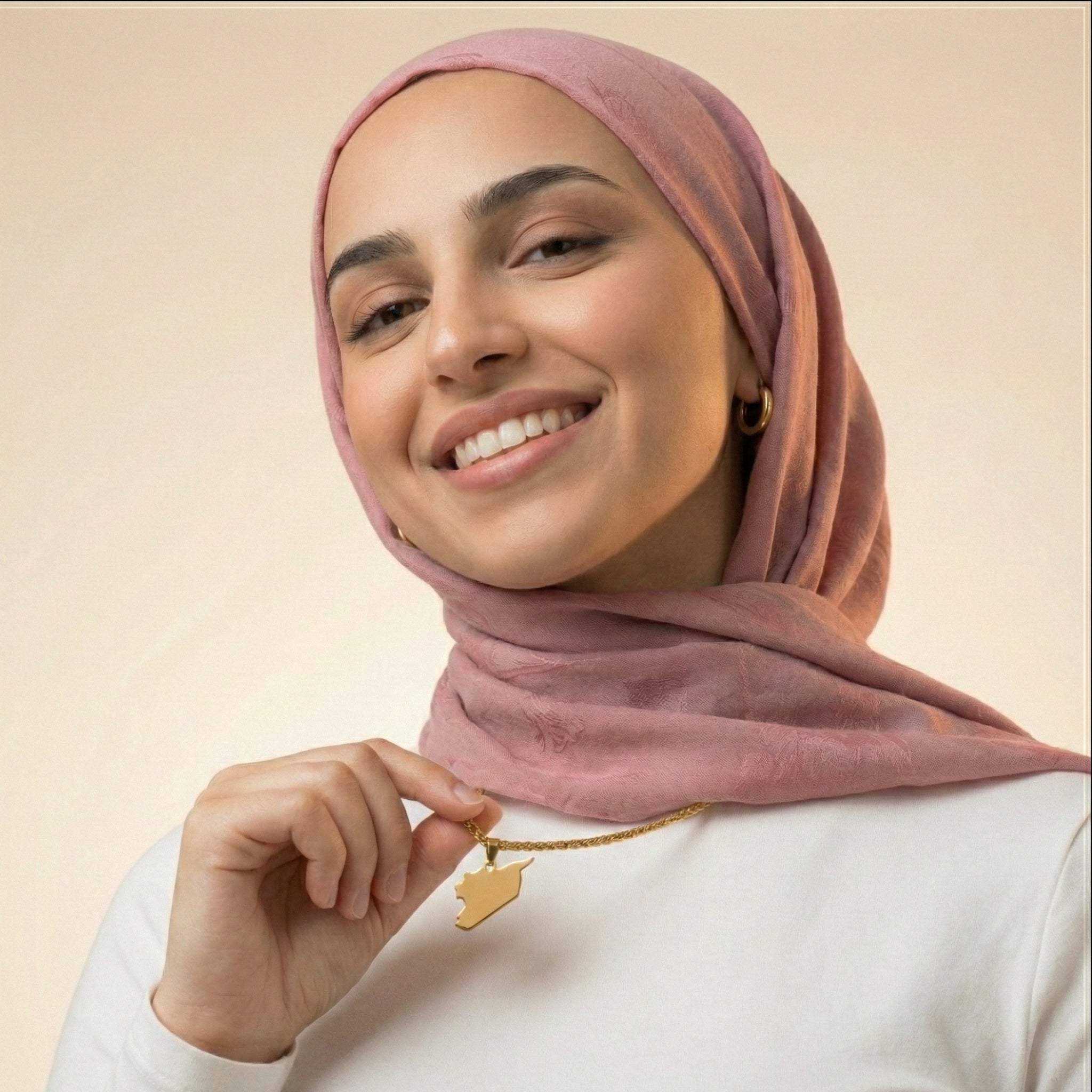 Woman wearing a pink hijab and holding a syria gold necklace with a star pendant against a beige background