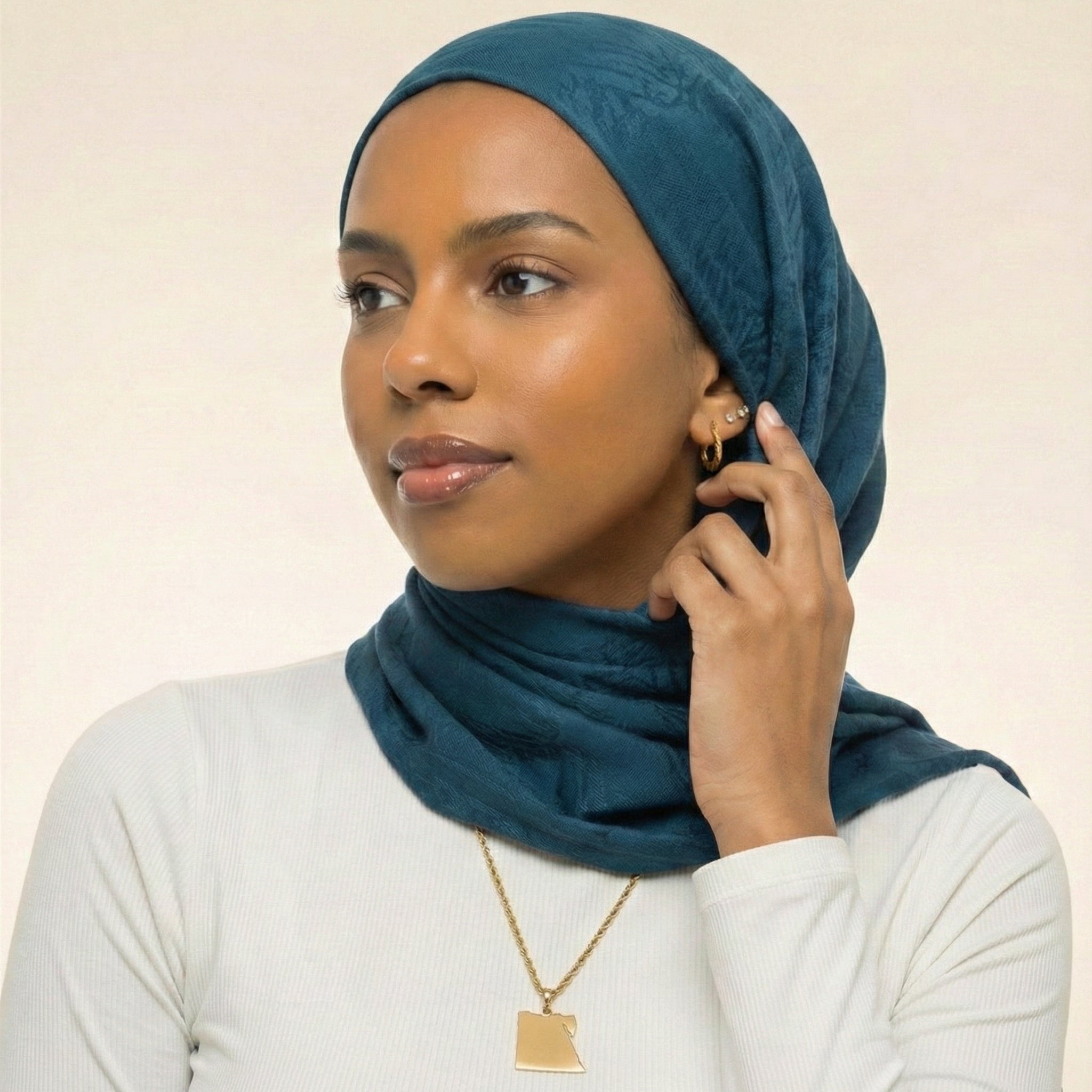 Woman wearing a teal hijab and white top against a beige background