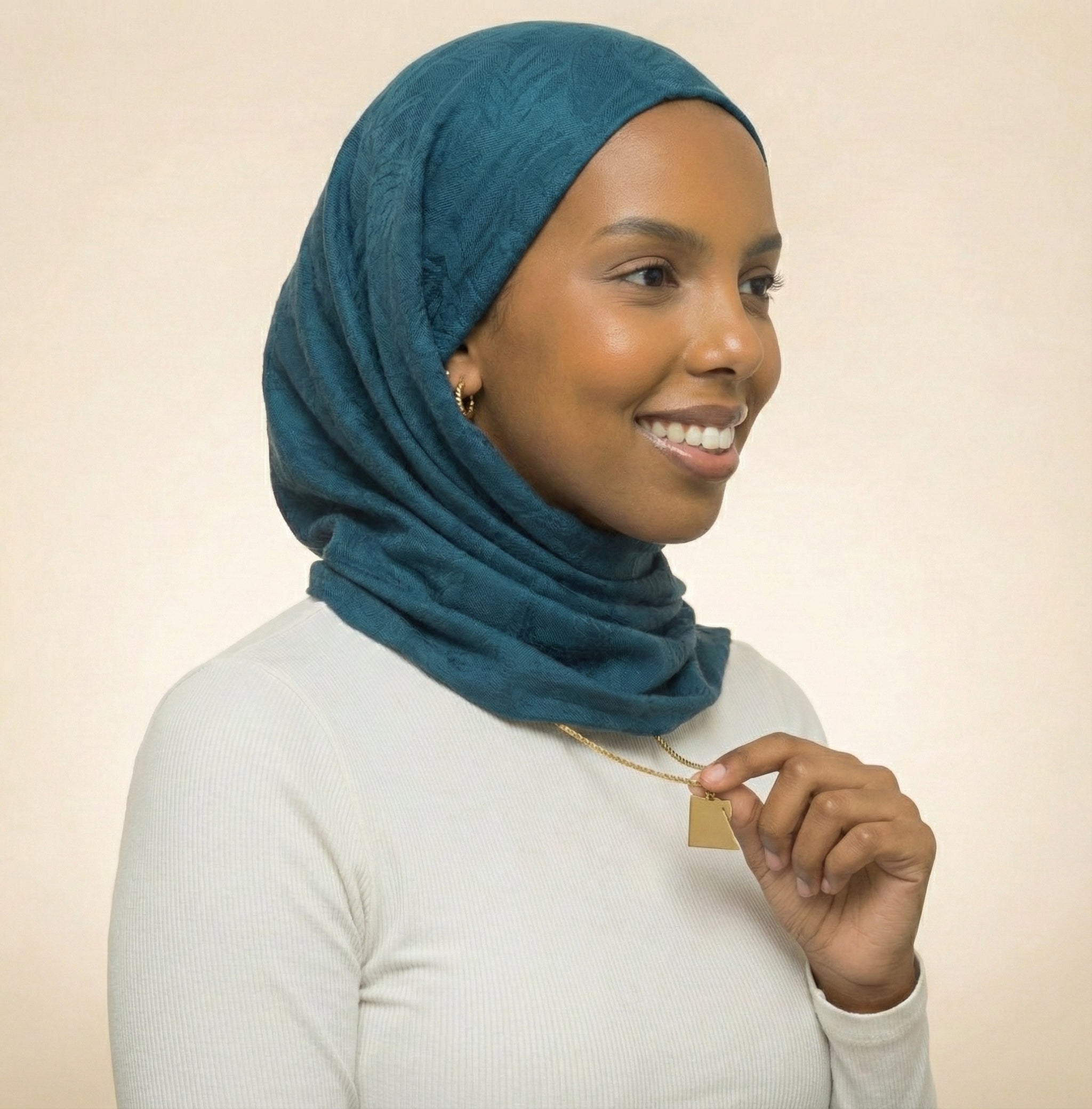 Woman wearing a teal hijab and holding a gold necklace against a beige background