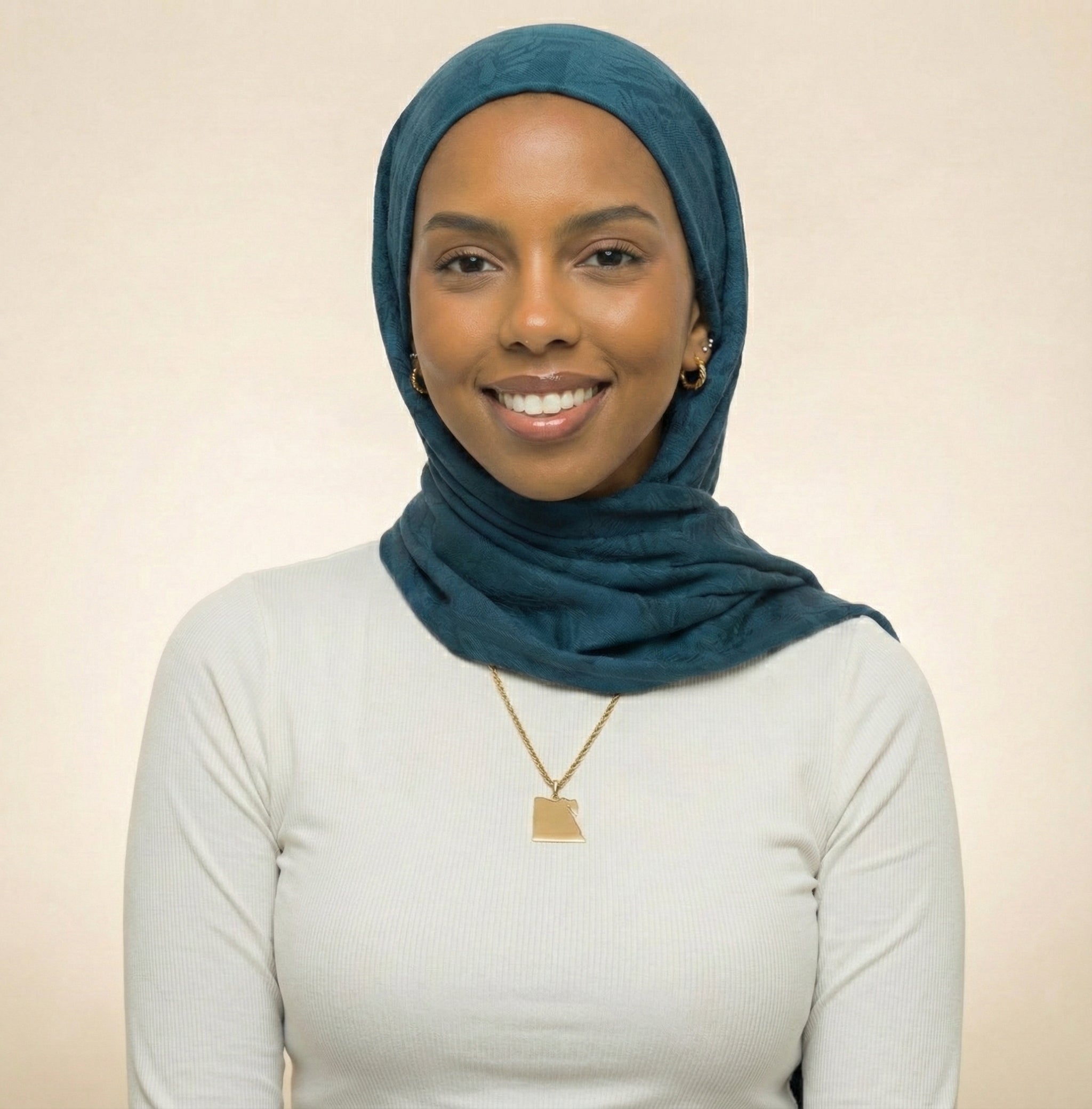 Woman wearing a teal hijab and gold necklace against a beige background