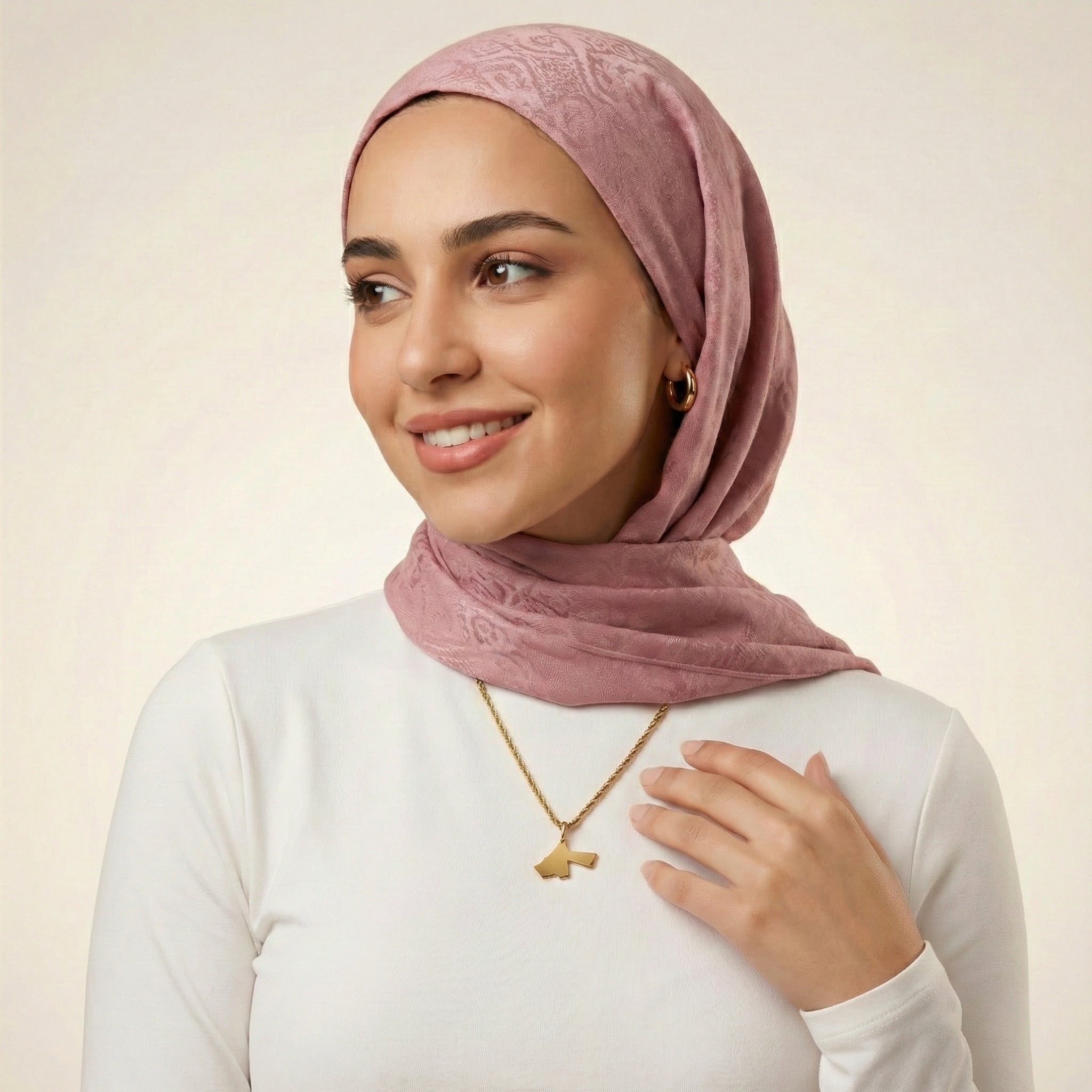 Woman wearing a pink hijab and gold necklace on a plain background