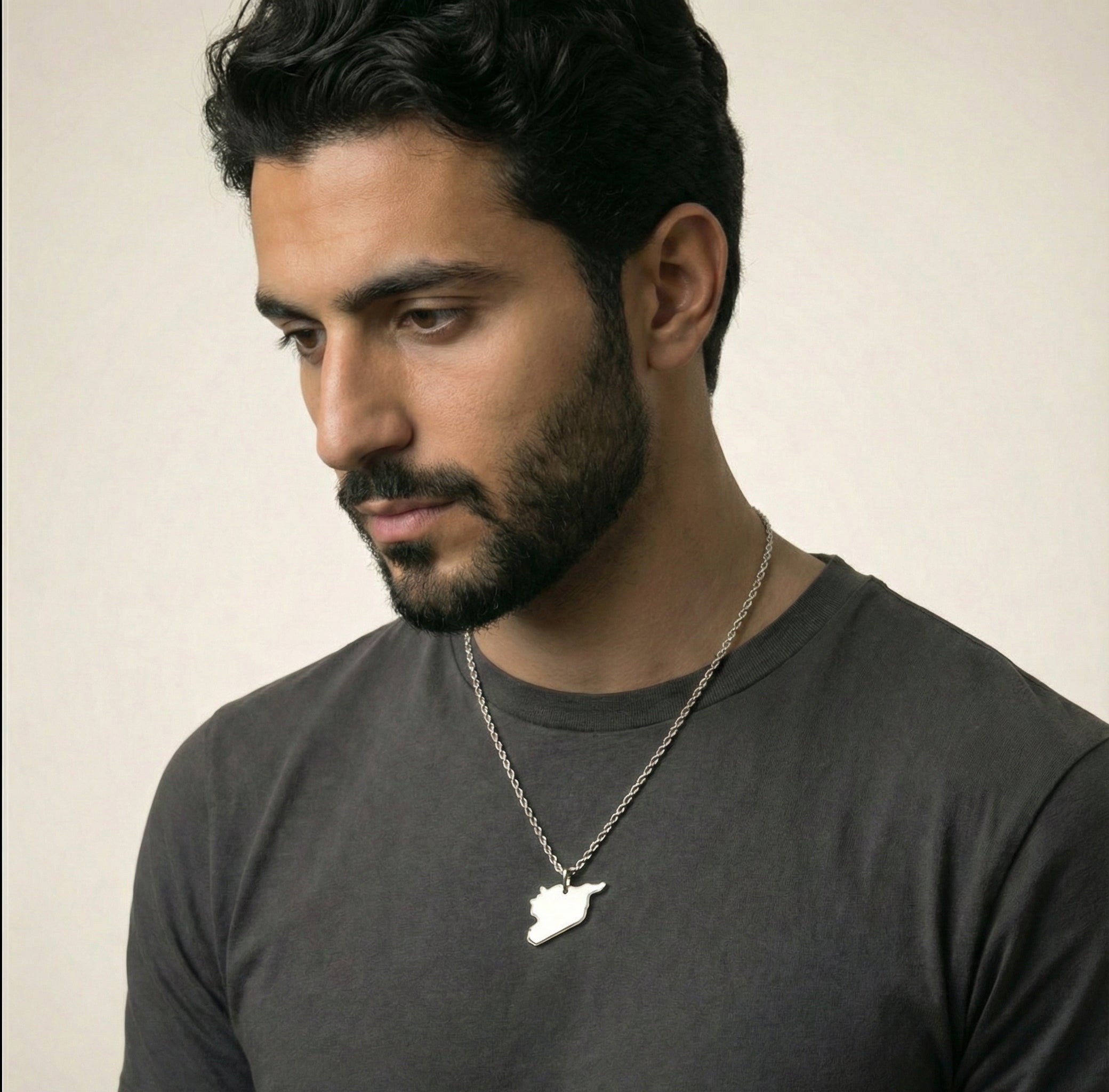 Man wearing a dark gray t-shirt with a necklace featuring a small syria pendant against a plain background