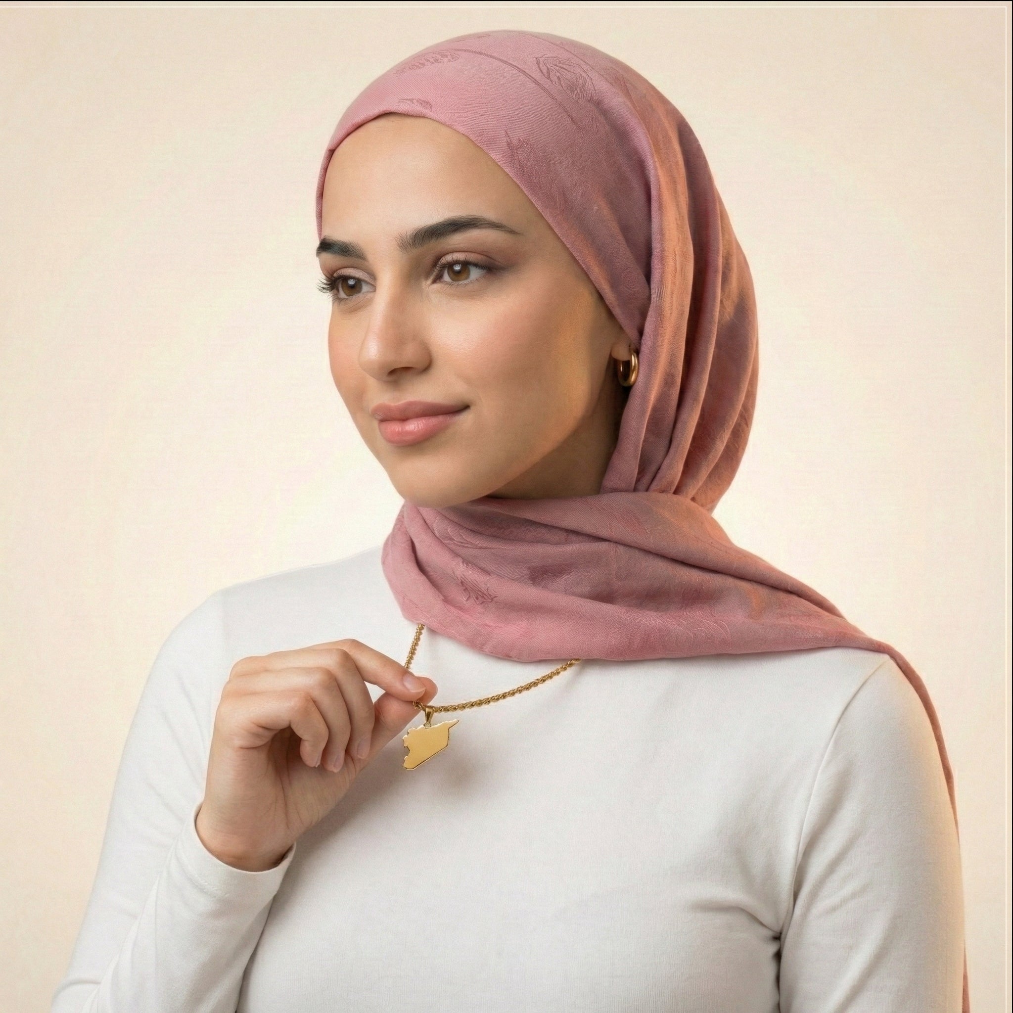 Woman wearing a pink hijab and holding a syria necklace against a beige background